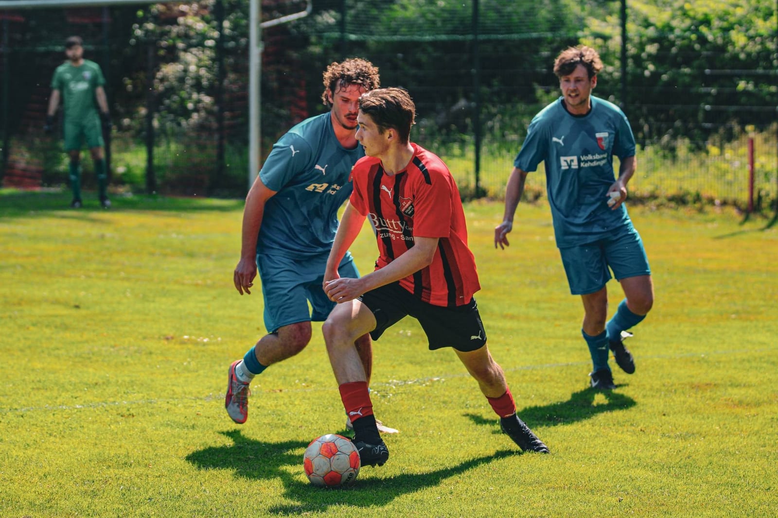 Spieler im rot-schwarzen Trikot dribbelt zwischen zwei Gegenspielern in Blau