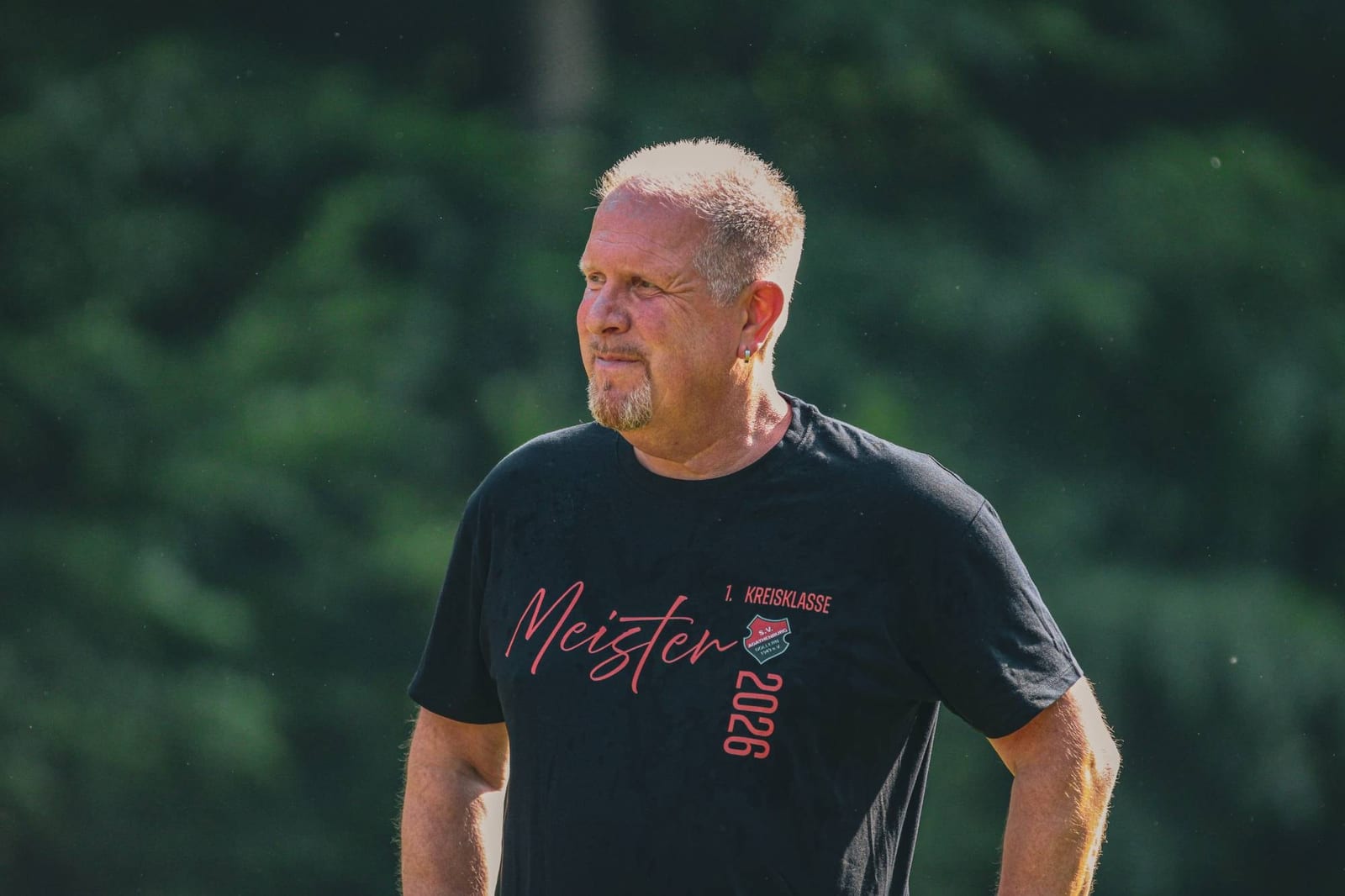 Grauhaariger Mann im Shirt mit der Aufschrift Meister 1. Kreisklasse 2026 schaut über den Platz