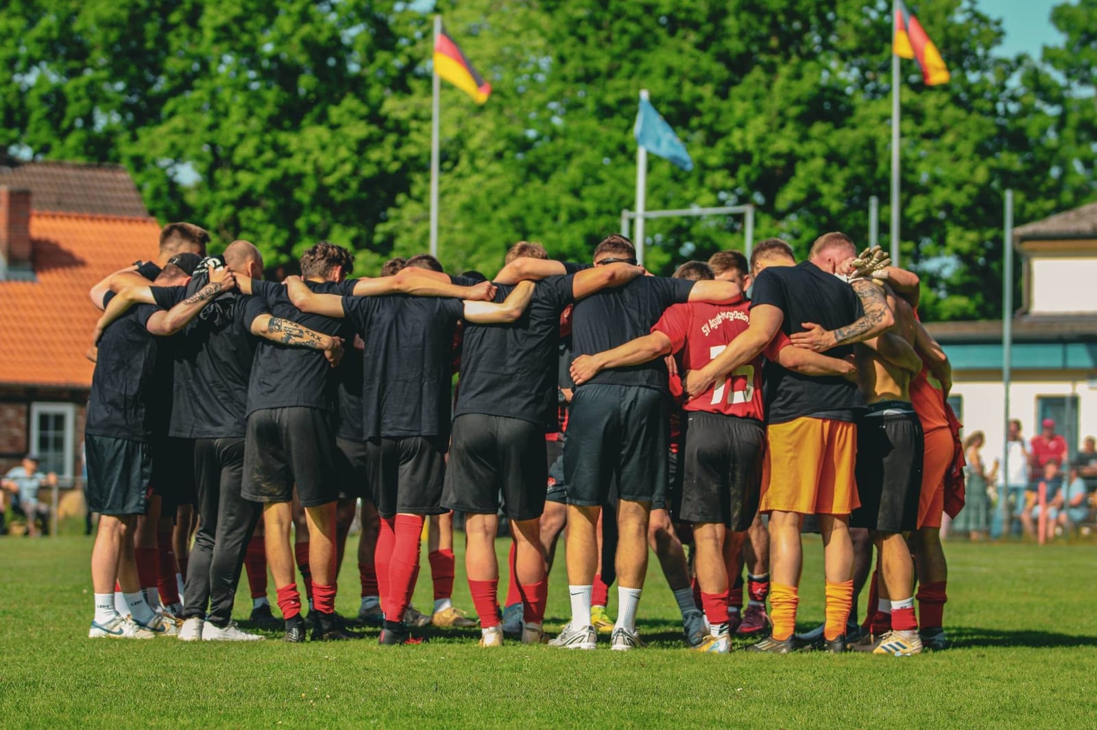 Die Mannschaft steht in schwarzen Meister-Shirts im Kreis auf dem Rasen, dahinter das Vereinsheim und Fahnen