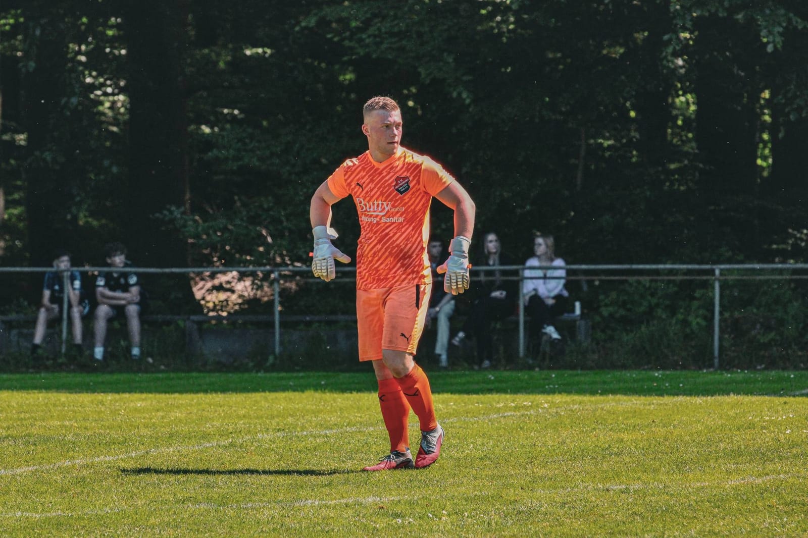Torwart im orangefarbenen Trikot kommt über den Platz, dahinter Bäume und Zuschauer am Zaun