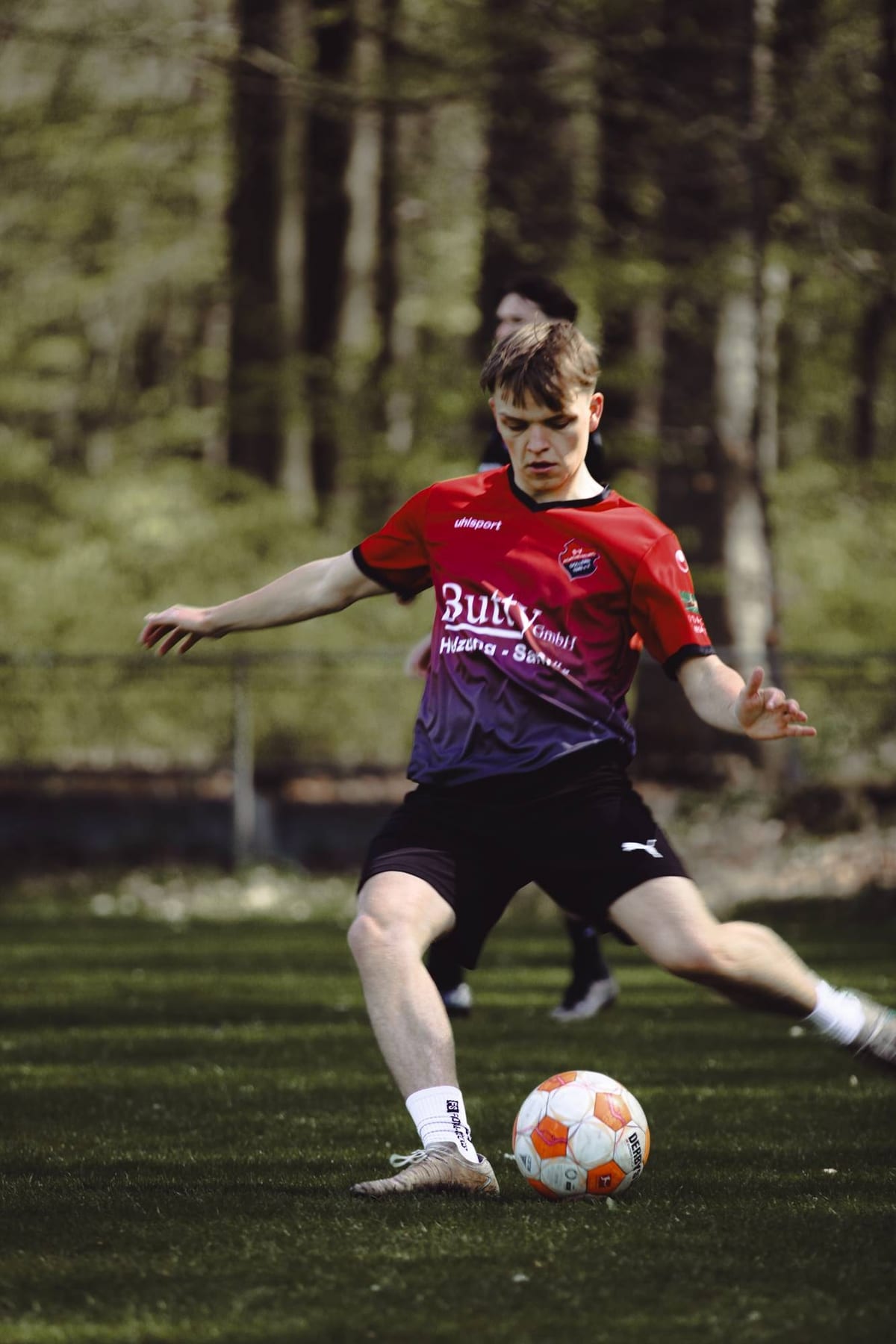 Spieler im Aufwärmshirt spielt den Ball, hinter ihm Kiefernwald im Frühlingslicht