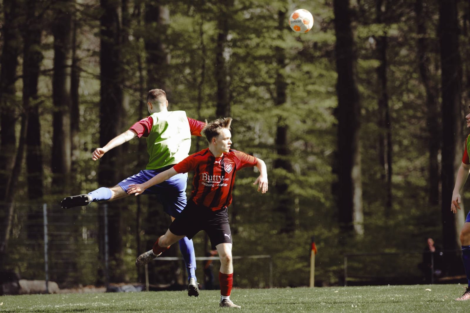 Zwei Spieler springen gleichzeitig zum hohen Ball, dahinter eine Reihe Kiefern