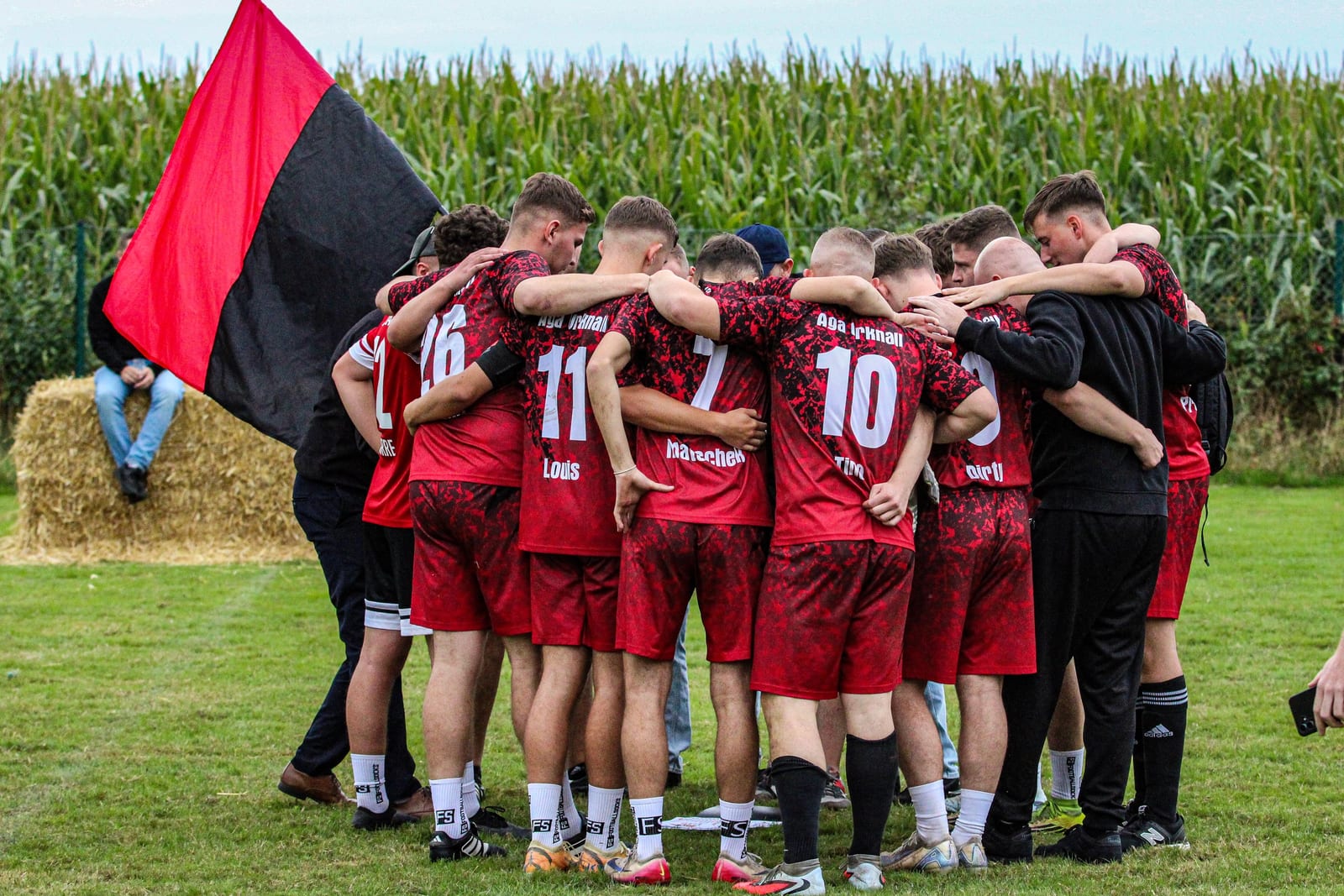 Mannschaft im roten Trikot steht vor dem Spiel im Kreis, daneben eine rot-schwarze Fahne