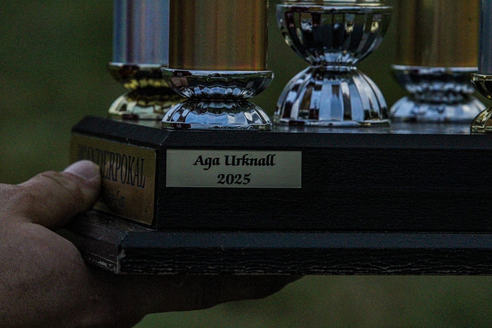 Eine Hand hält den Sockel eines Pokals mit der Gravur Aga Urknall 2025