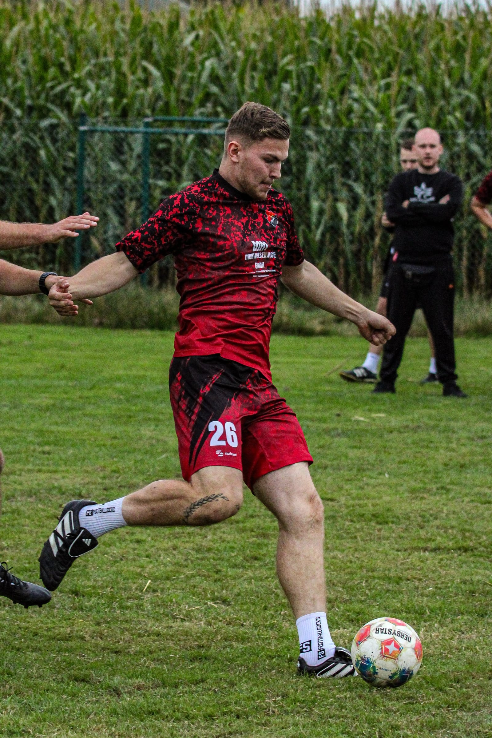 Spieler mit der Nummer 26 holt zum Schuss aus, dahinter ein Maisfeld