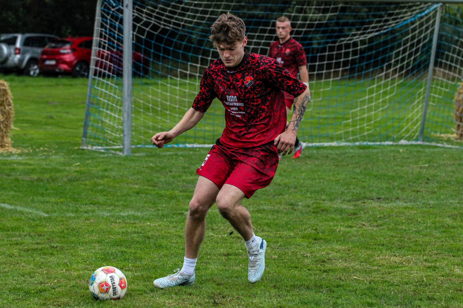 Spieler im rot-schwarzen Trikot nimmt den Ball vor dem Tor an, im Hintergrund der Torwart