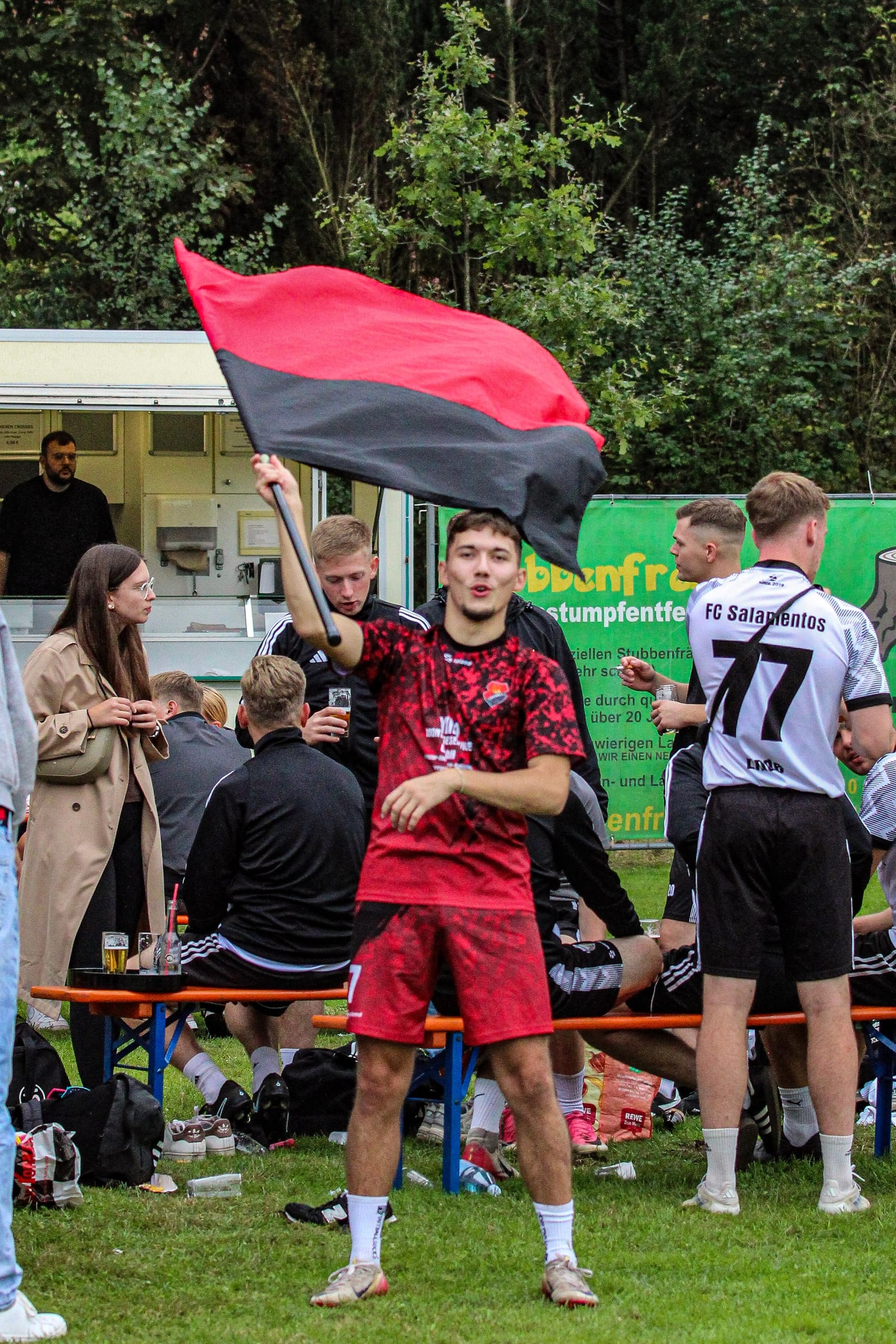 Spieler im roten Trikot schwenkt am Spielfeldrand eine rot-schwarze Fahne, hinter ihm Zuschauer an Bierbänken