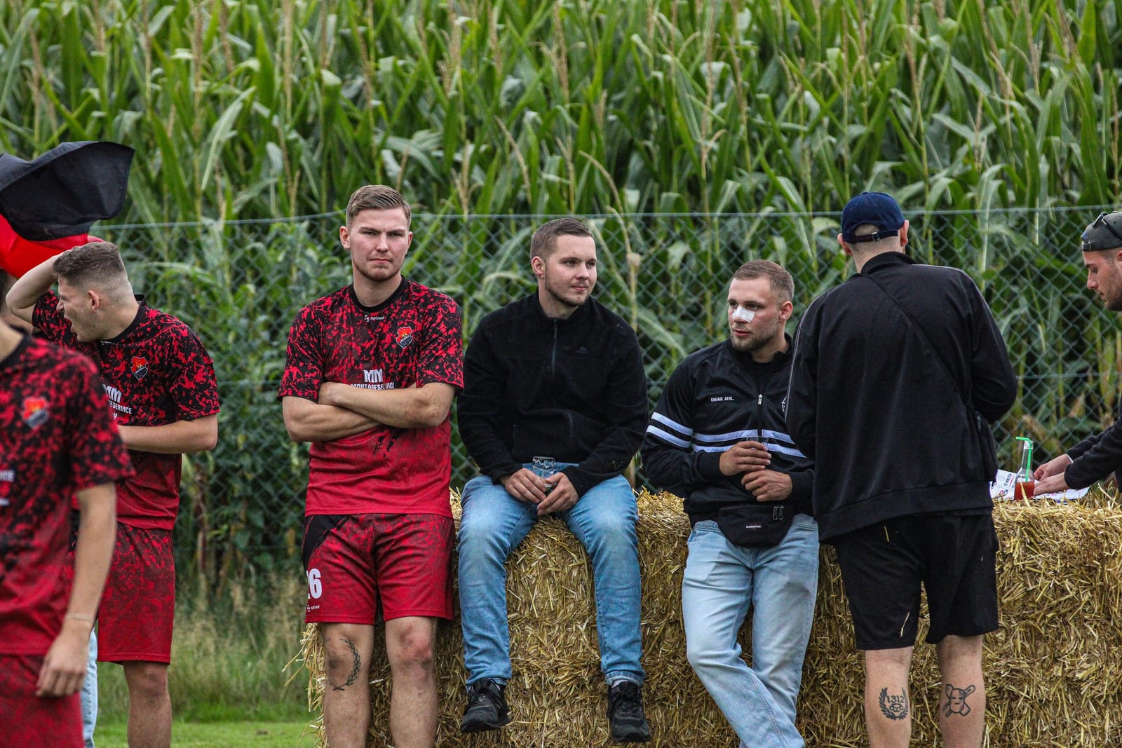 Spieler in roten Trikots und Begleiter stehen und sitzen vor dem Maisfeld an Strohballen