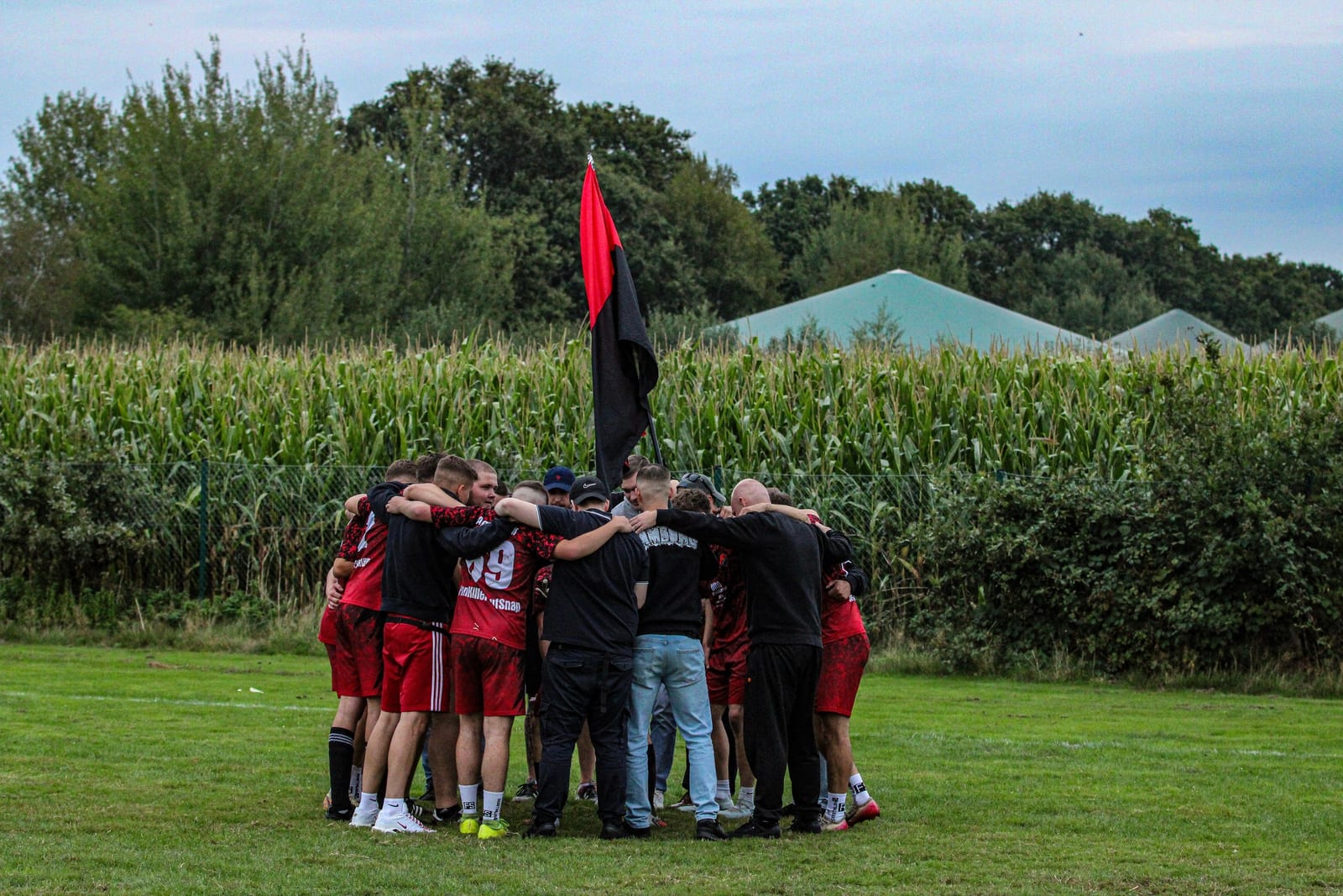 Die Mannschaft steht nach dem Sieg im Kreis auf dem Rasen, darüber eine rot-schwarze Fahne, dahinter das Maisfeld