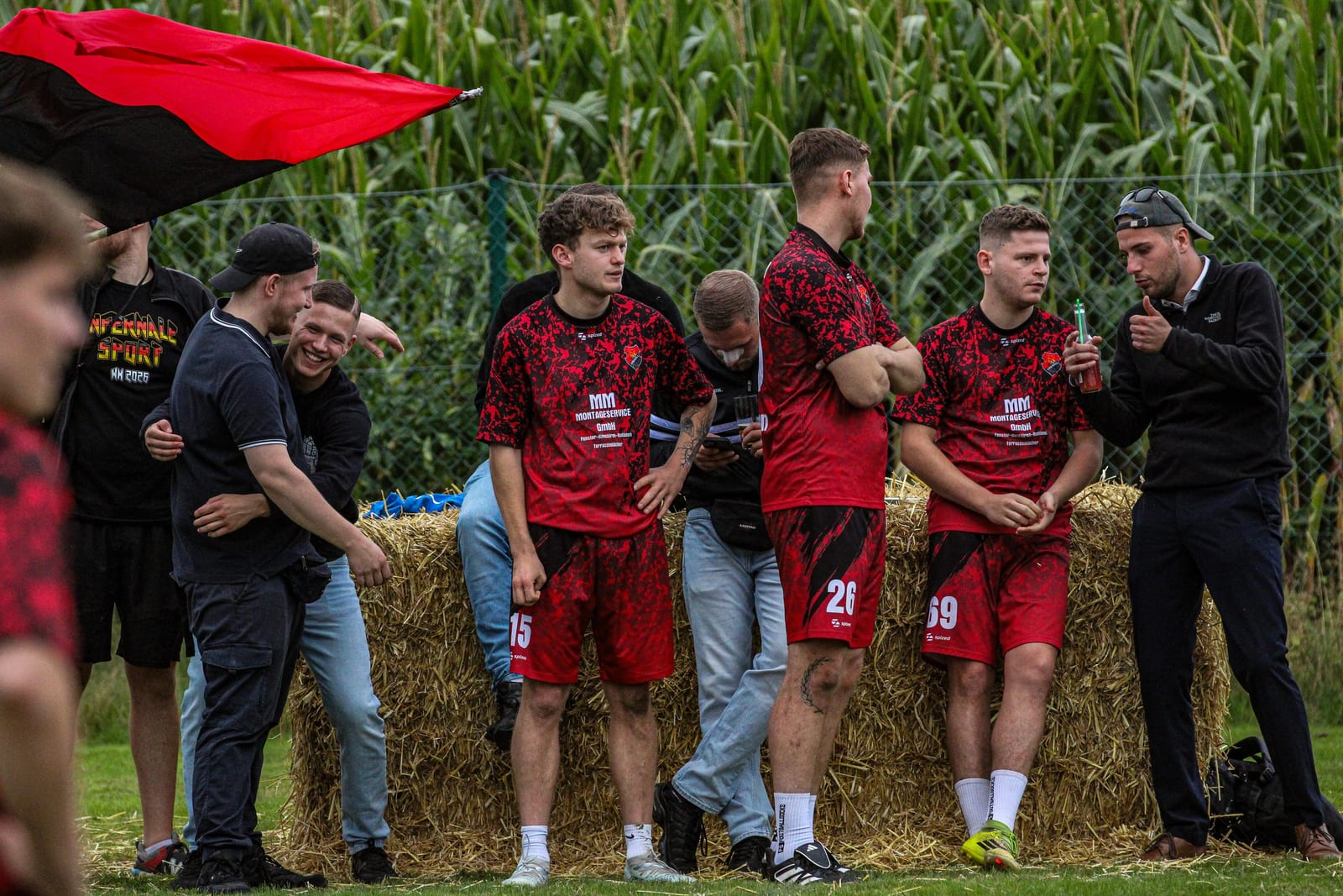 Spieler in roten Trikots warten an Strohballen vor dem Maisfeld auf ihr Spiel, eine rot-schwarze Fahne weht ins Bild