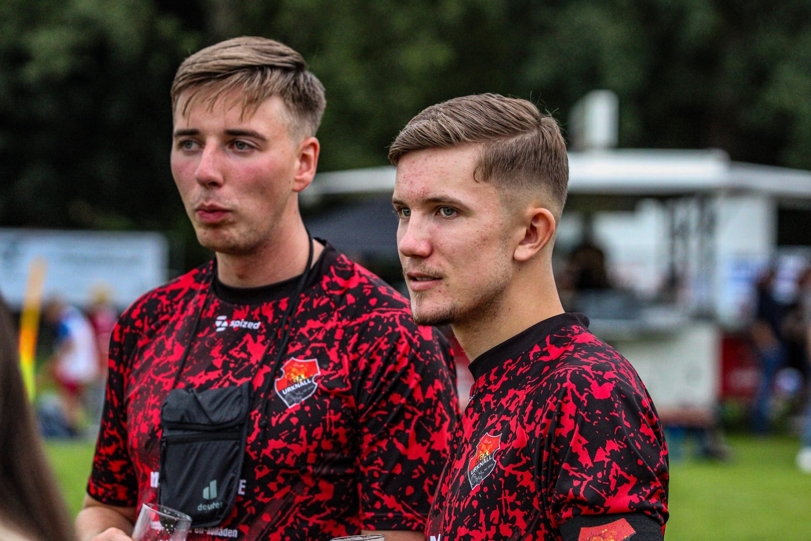 Zwei Spieler im rot-schwarzen Trikot verfolgen das Geschehen vom Spielfeldrand